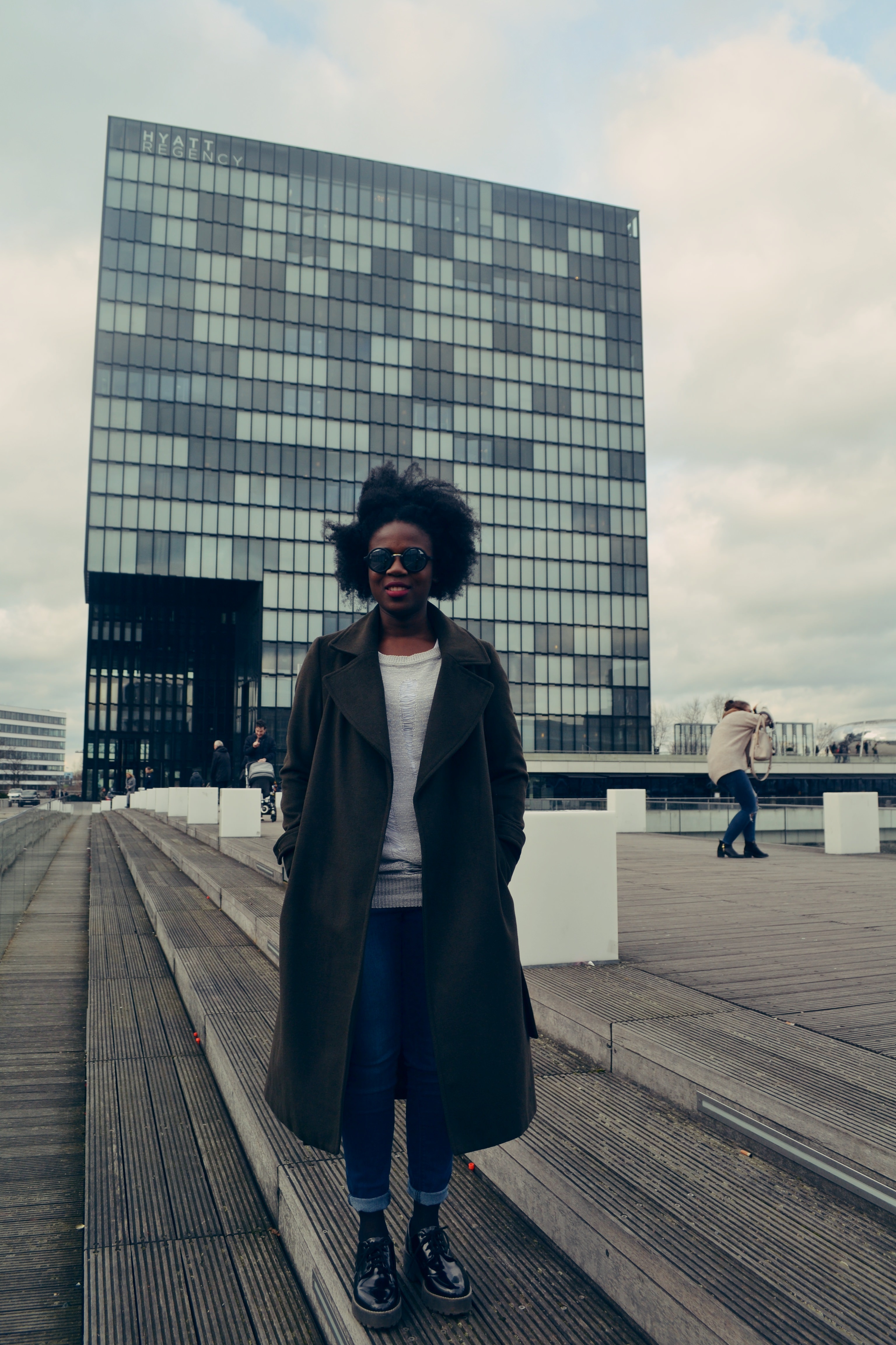 A woman stands in front of the Hyatt in Düsseldorf (the building looks like a square with black windows) in a khaki longline coat.