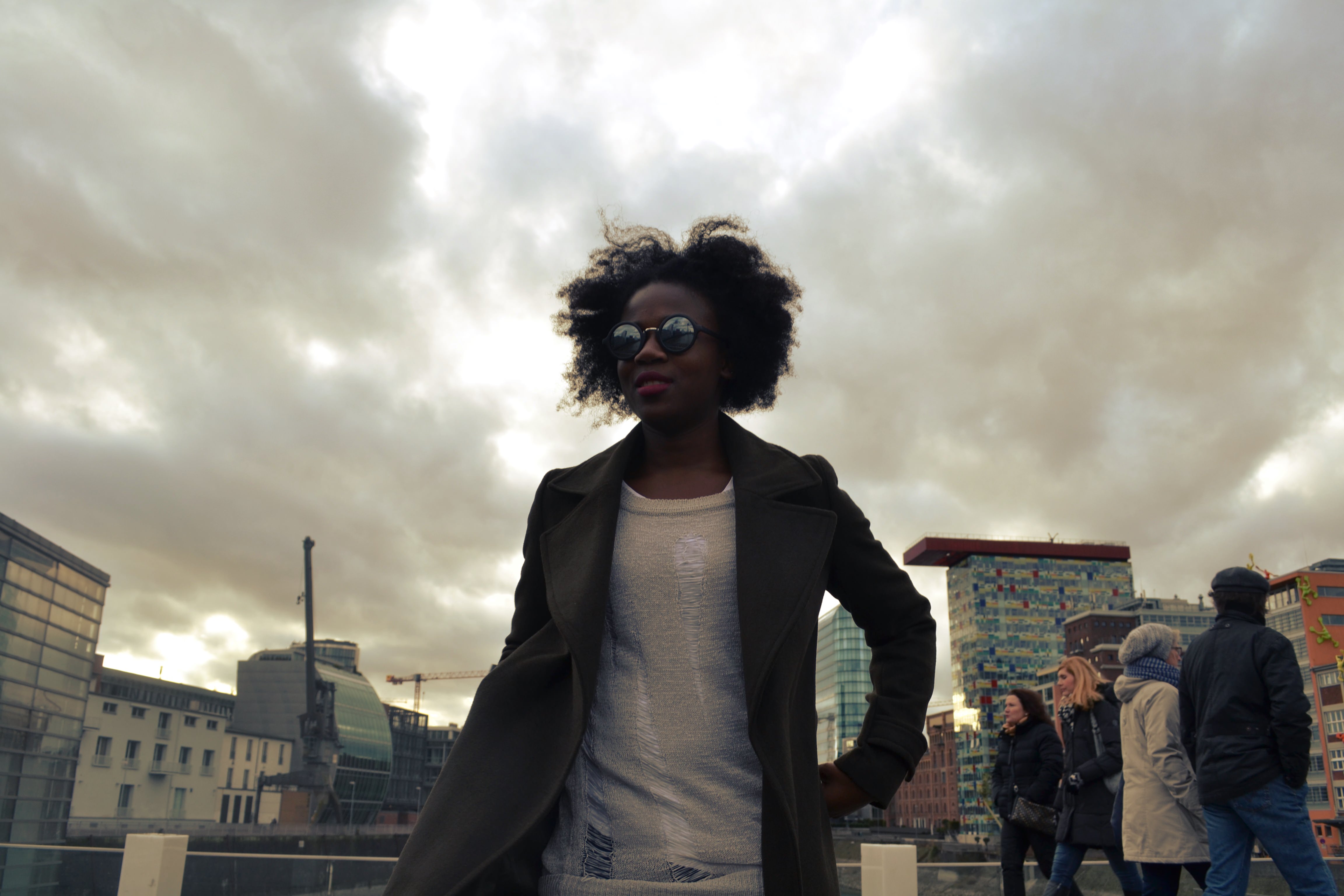 A woman stands in front of a cloudy sky with people walking in the background.