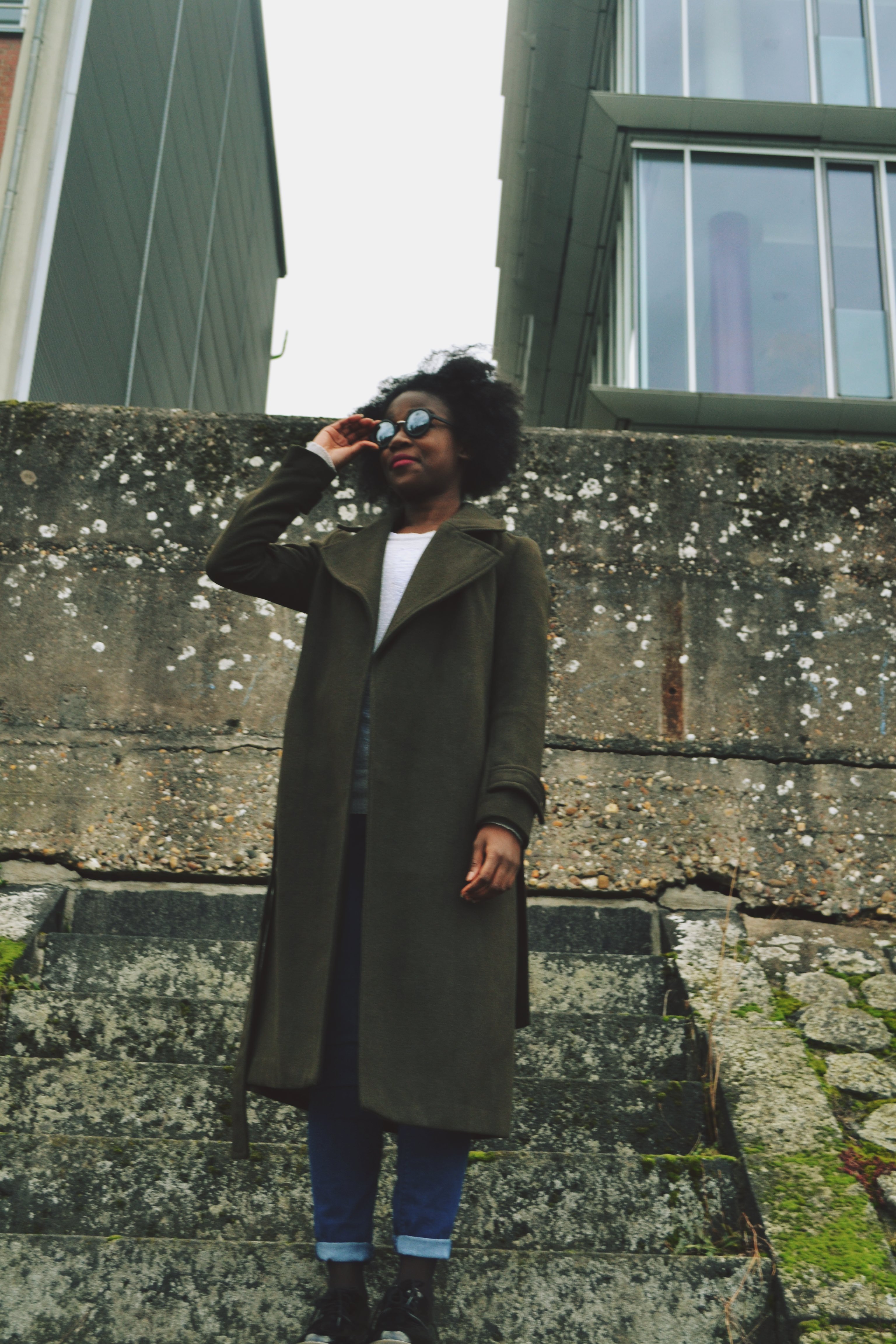 A woman with an afro is touching her sunglasses and smirks, looking off into the distance. She is wearing a khaki longline coat and jeans.