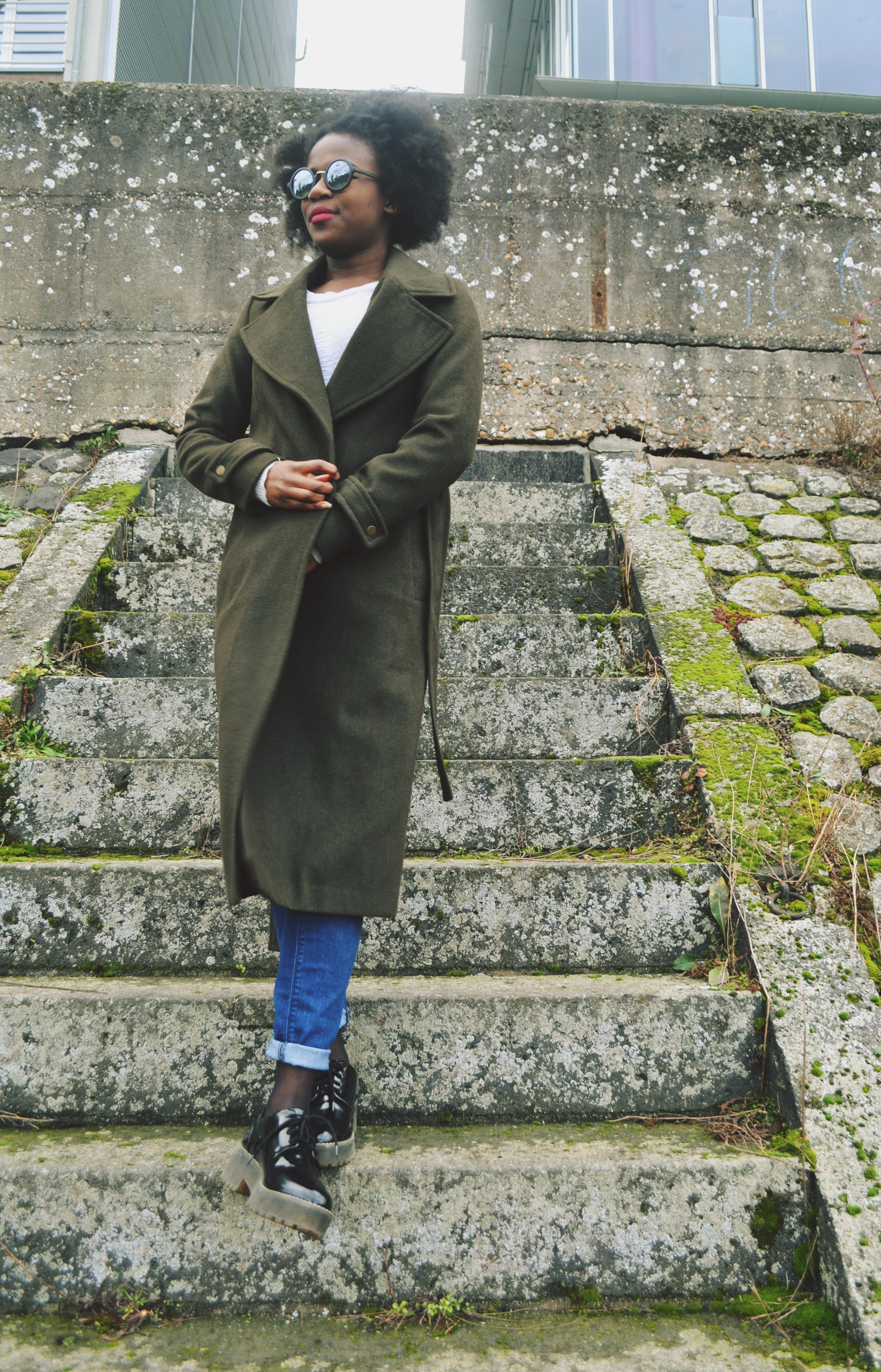 A woman walks down outdoor steps. She is wearing a longline khaki coat and holding it closed.