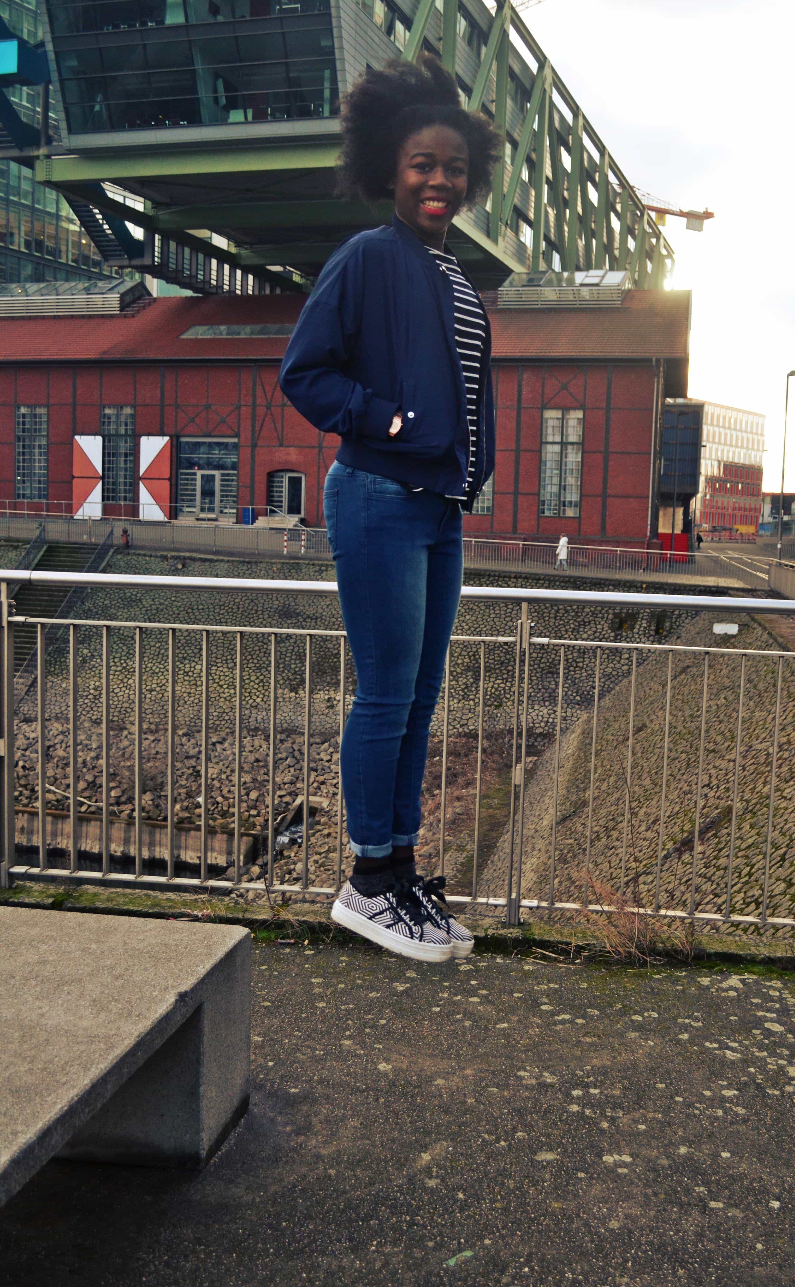 A woman with an afro wearing a navy blue bomber jacket, a black and white stripy t-shirt, blue skinny jeans and black and white stripy platform shoes jumps from a bench and smiles at the camera.